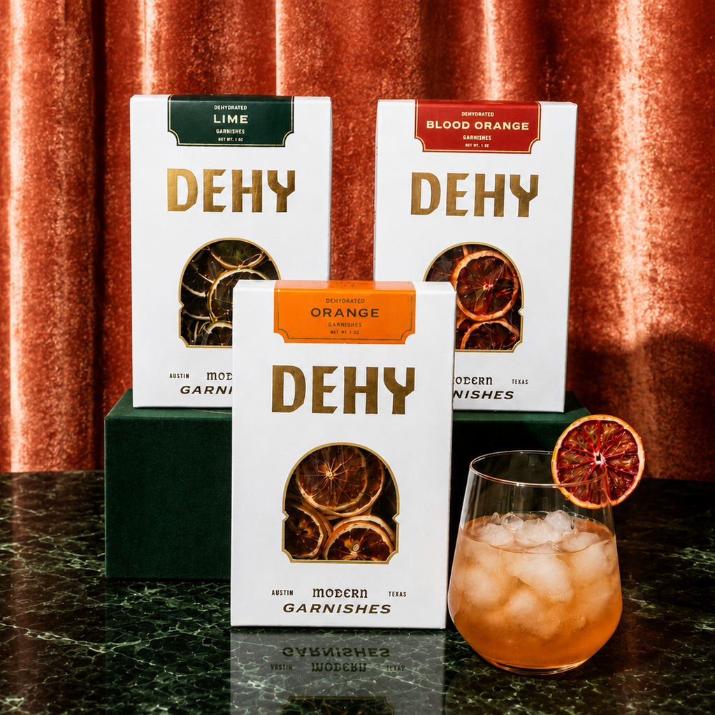 Three DEHY garnish boxes in Lime, Blood Orange, and Orange flavors arranged on a dark marble surface in front of a copper curtain backdrop, with a prepared orange cocktail and garnish slice beside them.
