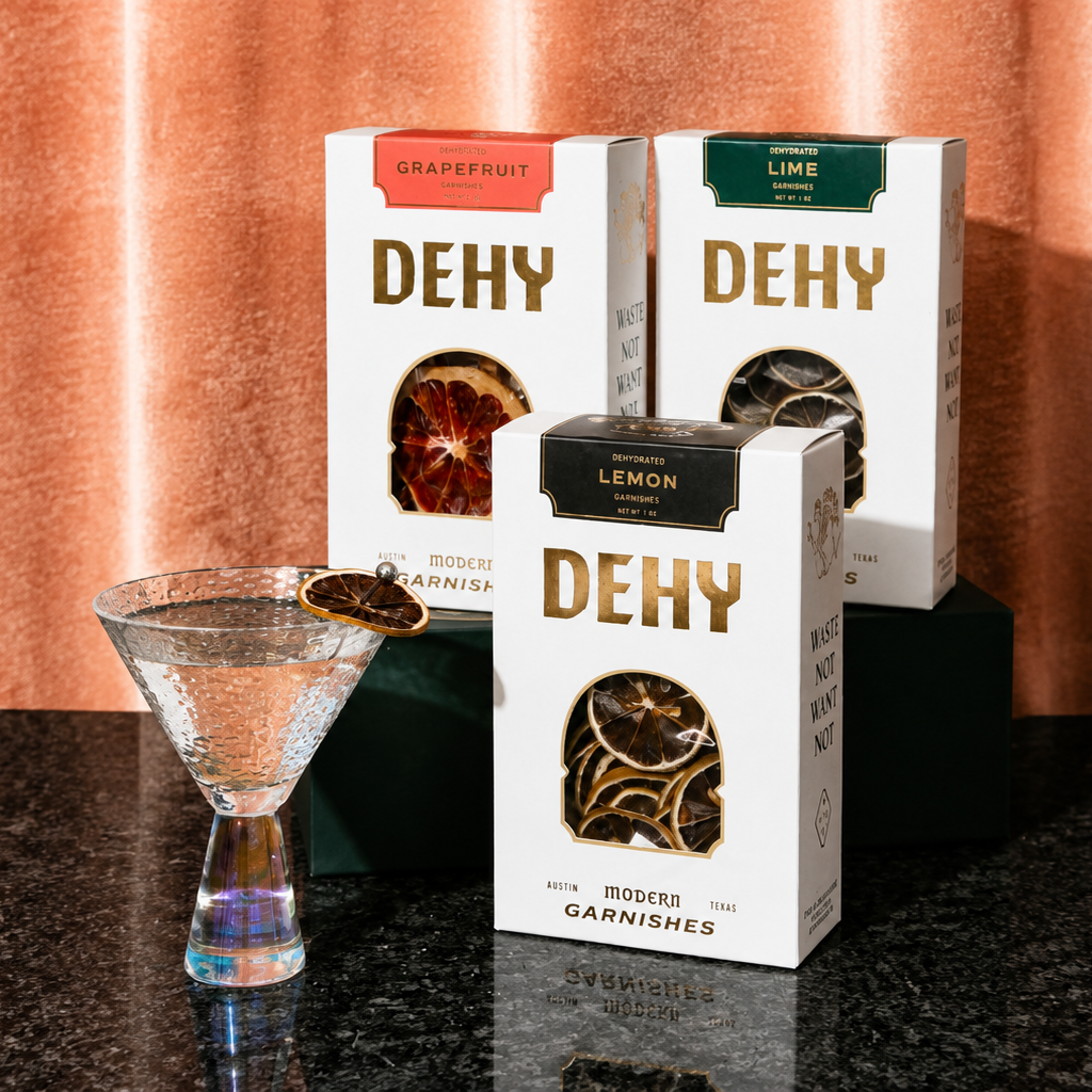 Three DEHY Garnish Boxes (Grapefruit, Lime, and Lemon) displayed on a black marble countertop with a textured copper background, alongside a martini glass garnished with a dried citrus peel.