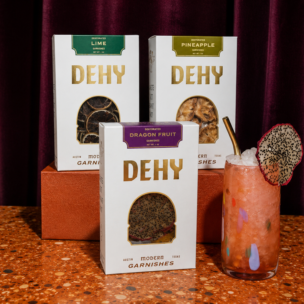 Three DEHY garnish boxes in white packaging with gold lettering are displayed on a terrazzo countertop in front of a dark purple curtain. A pink drink in a clear glass with a gold straw and a dragon fruit garnish sits to the right.